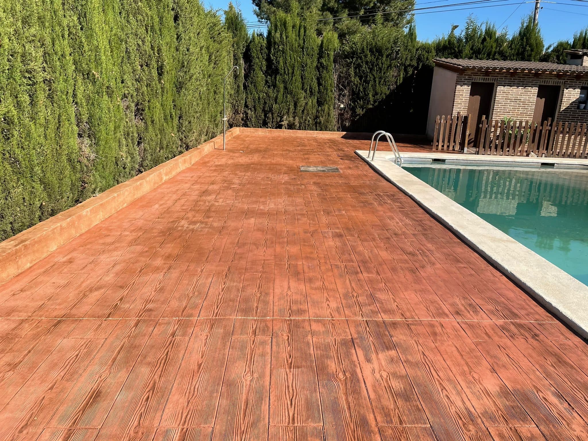 Contorno de piscina pavimentado en hormigón impreso con acabado madera en tono terracota, junto a un seto de cipreses