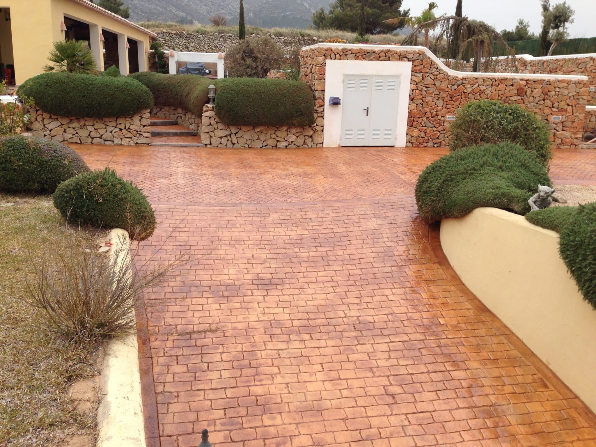 Acceso de vivienda en hormigón impreso con molde de ladrillo en espiga, junto a un muro de piedra